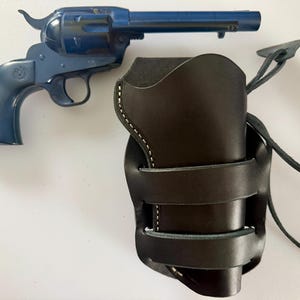 May include: A blue toy revolver and a black leather holster. The holster features white stitching and straps. The revolver is positioned beside the holster on a white surface.