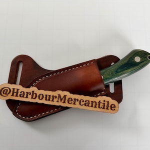 May include: A handcrafted knife with a green and black handle is partially sheathed in a brown leather holder. The holder is attached to a brown leather belt loop. A wooden tag with the text "@HarbourMercantile" is in front.
