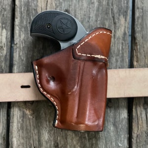Può includere: Un fodero in pelle marrone con cuciture bianche per una piccola pistola. Il fodero è progettato per essere indossato su una cintura.