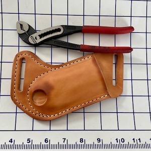 Könnte beinhalten: Eine schwarze und rote Zange mit einem Leder-Werkzeughalter. Die Zange hat einen silbernen Kopf und rote Griffe. Der Werkzeughalter ist hellbraun und hat weiße Nähte an den Kanten.