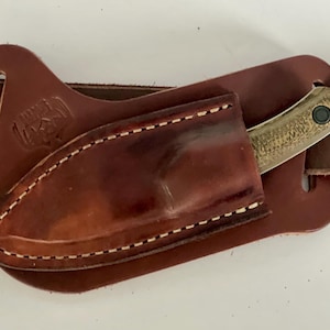 May include: A brown leather knife sheath with a small knife inserted. The sheath is attached to a brown belt. The knife has a textured handle and a silver blade. The sheath is stitched with white thread.