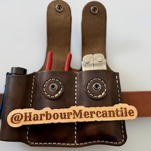 May include: A brown leather tool belt with a multi-tool, pliers, and a flashlight. The belt has a wooden tag that reads "@HarbourMercantile". The belt is designed to hold tools and accessories.