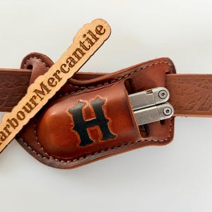 May include: A brown leather belt holster featuring a black letter H embossed on the front, holding a silver multi-tool. The holster is threaded onto a matching brown leather belt with a textured pattern. A wooden tag reads @HarbourMercantile.