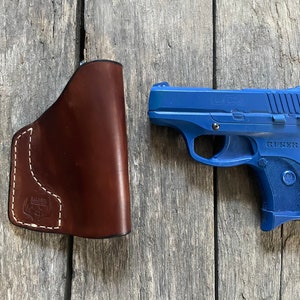 Może przedstawiać: Niebieski pistolet Ruger LCP z brązową sk&oacute;rzaną kaburą. Pistolet ma czarną lufę i srebrny zamek. Kabura wykonana jest ze sk&oacute;ry brązowej i ma białe szwy.