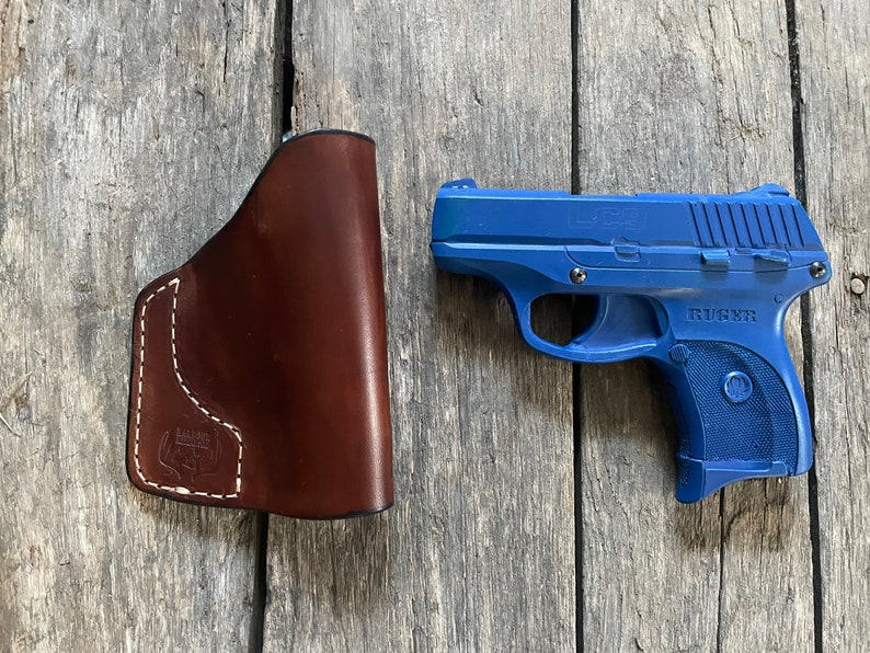 Może przedstawiać: Niebieski pistolet Ruger LCP z brązową sk&oacute;rzaną kaburą. Pistolet ma czarną lufę i srebrny zamek. Kabura wykonana jest ze sk&oacute;ry brązowej i ma białe szwy.