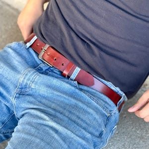 May include: A brown leather belt with a silver buckle is worn with blue jeans. The belt has a classic design with a rectangular buckle and a series of holes for adjustment. The belt is worn with a dark gray t-shirt.
