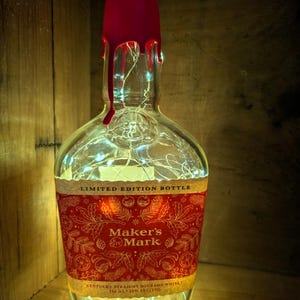 May include: A clear glass Maker's Mark bourbon bottle with a red wax seal and a decorative red and gold label. The bottle is lit with fairy lights inside. The label reads "LIMITED EDITION BOTTLE" and "Maker's Mark".