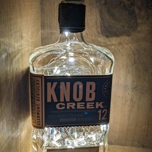 May include: A clear glass Knob Creek 12 Kentucky Straight Bourbon Whiskey bottle with a black label and cap. The label reads "Knob Creek" and "12". The bottle is illuminated with small white lights.