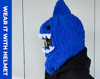 Op de afbeelding: Blauwe gebreide balaclava met een witte grijns met tanden en zwarte goggles. De balaclava wordt gedragen over een zwart jack. "WEAR IT WITH HELMET" staat links op de afbeelding.