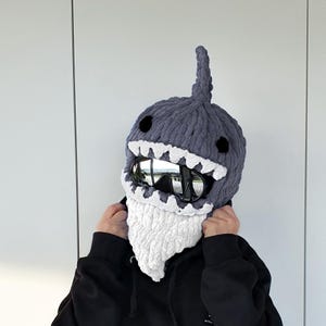 May include: A gray knitted shark hat with white teeth and black eyes. The hat has a fin on top and a white beard. The hat is worn with black goggles.