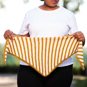 May include: A knitted shawl with a striped pattern in mustard yellow and cream. The triangular shawl is held up, showcasing the design. The background is blurred, with natural light and greenery.