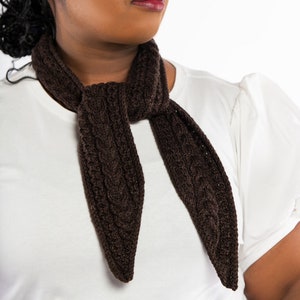 May include: A woman wearing a white shirt with a brown knitted scarf. The scarf has a cable knit pattern.