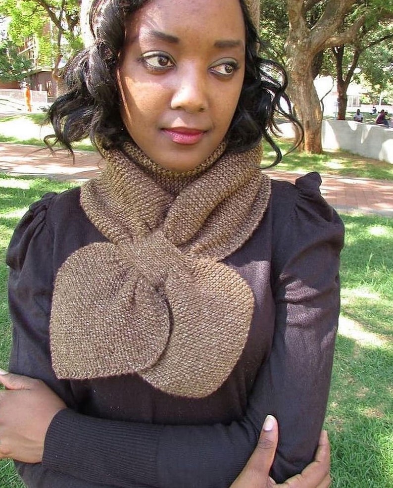May include: A brown knitted scarf with a large bow in the front. The scarf is worn by a person with dark hair and brown eyes.