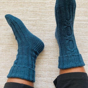 May include: A pair of dark teal knitted socks featuring a vertical ribbed pattern and a central cable knit design running down the front. The socks have a folded cuff at the top and are crafted from a textured yarn, extending to the mid-calf area.