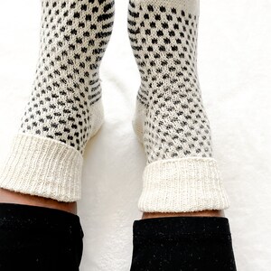 Puede incluir: Un par de calcetines blancos de punto con un patr&oacute;n geom&eacute;trico negro. Los calcetines est&aacute;n doblados en la parte superior, revelando un pu&ntilde;o de punto blanco.