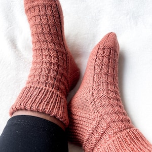 Pode incluir: Um par de meias de malha cor coral com um padrão geométrico texturizado no pé e punho canelado. As meias são exibidas em uma superfície branca, mostrando o trabalho de malha detalhado e a cor quente.