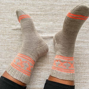 May include: A pair of knitted socks in a neutral beige color with orange accents. The socks feature an orange geometric pattern around the ankle and stripes near the toes. The socks are worn with black pants.