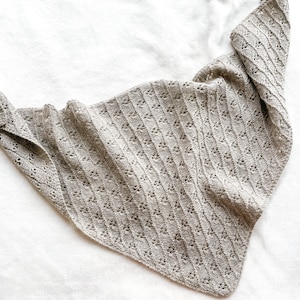 May include: A light brown knitted shawl with a delicate openwork pattern. The shawl is folded in half and laid flat on a white surface.