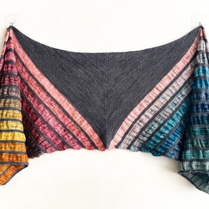 Pode incluir: Um xale triangular com um degradê de cores, incluindo amarelo, laranja, vermelho, rosa, azul e verde. O xale é feito de um fio macio e texturizado e tem uma borda franjada.