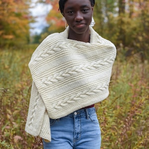 May include: A woman wearing a white cable knit sweater with a brown and white pattern. She is standing in a field of tall grass with fall foliage in the background.
