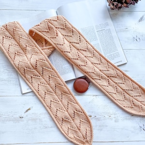 Peut inclure: Une écharpe en tricot de couleur pêche avec un motif en chevron. L'écharpe est posée sur une surface en bois blanc.