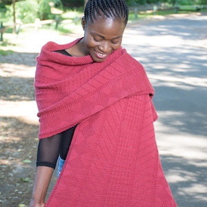May include: A woman wearing a red knitted shawl with a geometric pattern. The shawl is draped over her shoulders and she is holding the edge of the shawl in her hand.
