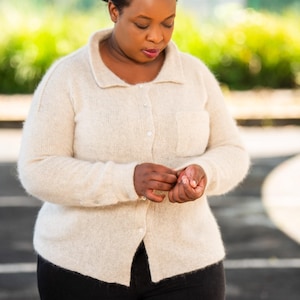 May include: A woman wearing a cream-coloured cardigan with a button-up front and black trousers. She is adjusting the sleeve of the cardigan.