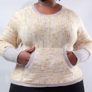 Puede incluir: Un suéter de color crema con un patrón moteado amarillo y un gran bolsillo en la parte delantera. El suéter tiene un ribete gris alrededor del cuello y el bolsillo.