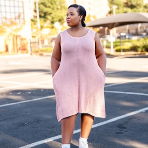 May include: A sleeveless, light pink knit dress with a scoop neckline and two front pockets. The dress has a vertical seam down the front and is paired with white high-top sneakers. The person is standing on an asphalt surface.