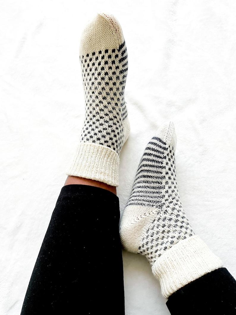 Puede incluir: Un par de calcetines blancos de punto con un patr&oacute;n geom&eacute;trico blanco y negro. Los calcetines se llevan puestos en los pies de una persona con pantalones negros.