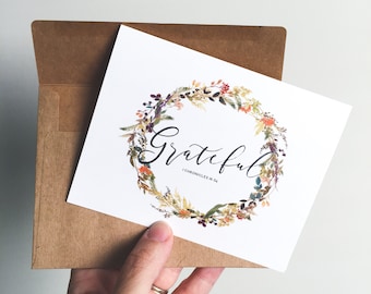 May include: A white card with a watercolor wreath of fall leaves and flowers. The word "Grateful" is written in cursive script inside the wreath. The text "1 Chronicles 16:34" is written below the word "Grateful". The card is in a brown envelope.