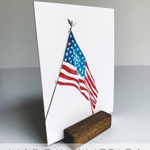May include: A watercolor painting of an American flag waving in the wind. The flag is mounted on a wooden stand with the words "MADE IN AMERICA" printed below the image.