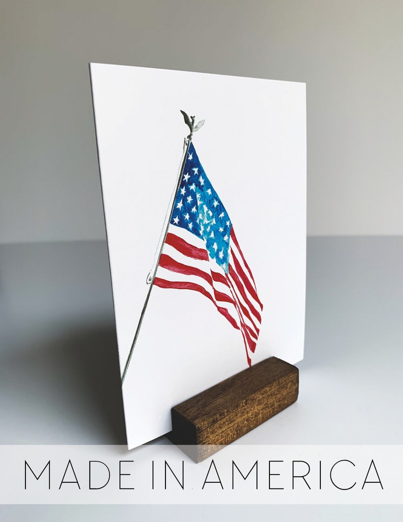 May include: A watercolor painting of an American flag waving in the wind. The flag is mounted on a wooden stand with the words "MADE IN AMERICA" printed below the image.