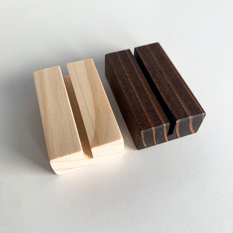 May include: Two wooden card holders, one natural wood and one dark brown stained wood. Both have a slot for holding a card or sign.