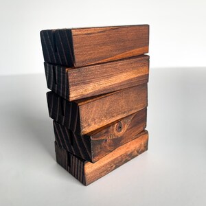 May include: A stack of six wooden blocks, each with a dark brown stain and a slightly different grain pattern. The blocks are stacked in a staggered pattern, creating a unique and rustic look.