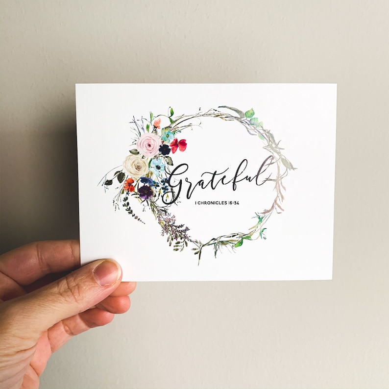 May include: A white card with a watercolor wreath of flowers in shades of pink, red, blue, and green. The word "Grateful" is written in black script inside the wreath. The text "1 Chronicles 16:34" is written in black below the word "Grateful".