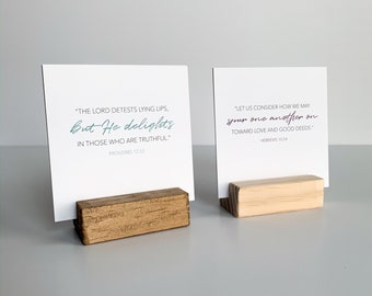 May include: Two wooden card holders with inspirational quotes printed on white cards. The quote on the left card reads "The Lord detests lying lips, But He delights in those who are truthful." Proverbs 12:22. The quote on the right card reads "Let us consider how we may spur one another on toward love and good deeds." Hebrews 10:24.