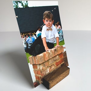 May include: A young boy in a white shirt and black pants sits on a brick wall. He is wearing a brown bow tie and has a serious expression on his face. The photo is printed on a white background and is displayed in a wooden photo stand.
