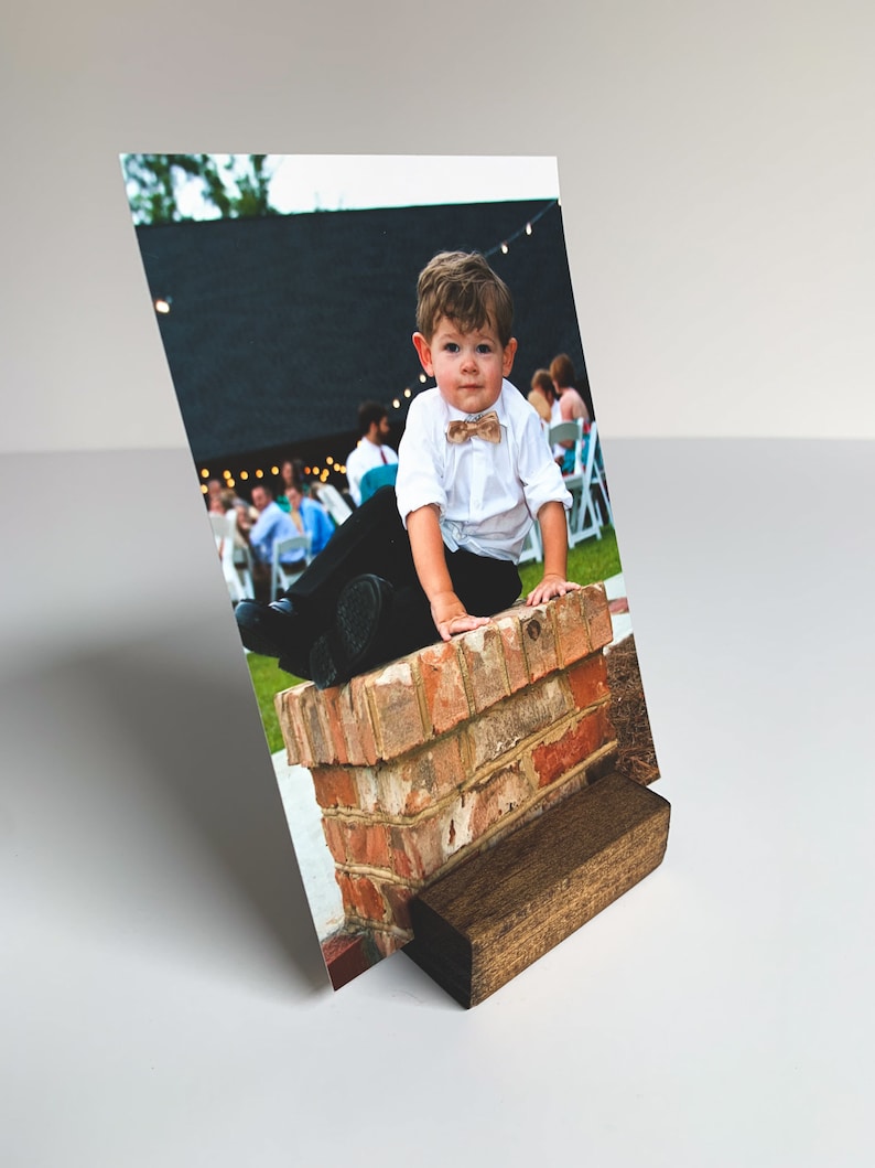 May include: A young boy in a white shirt and black pants sits on a brick wall. He is wearing a brown bow tie and has a serious expression on his face. The photo is printed on a white background and is displayed in a wooden photo stand.