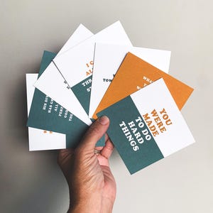 Op de afbeelding: Een hand houdt een waaier van vierkante kaarten met inspirerende citaten vast. De kaarten hebben witte, teal en oranje achtergronden met tekst in complementaire kleuren. Eén kaart luidt: "YOU WERE MADE TO DO HARD THINGS."