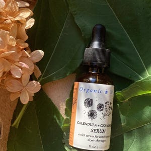 Może przedstawiać: Brązowa szklana butelka z czarną pipetą, oznaczona napisem "Organic & W" i "Calendula + Chamomile Serum". Etykieta zawiera ilustracje kwiatowe i tekst. Butelka jest umieszczona na tle zielonych liści i suszonych kwiat&oacute;w.