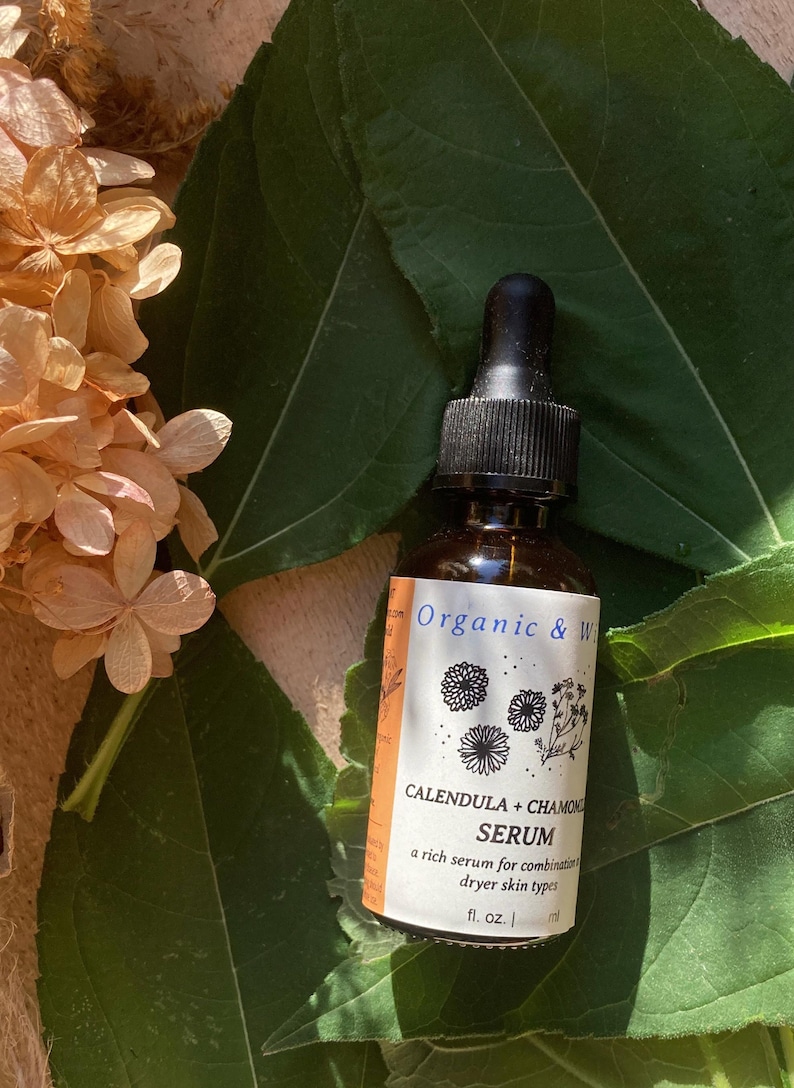 Może przedstawiać: Brązowa szklana butelka z czarną pipetą, oznaczona napisem "Organic & W" i "Calendula + Chamomile Serum". Etykieta zawiera ilustracje kwiatowe i tekst. Butelka jest umieszczona na tle zielonych liści i suszonych kwiat&oacute;w.