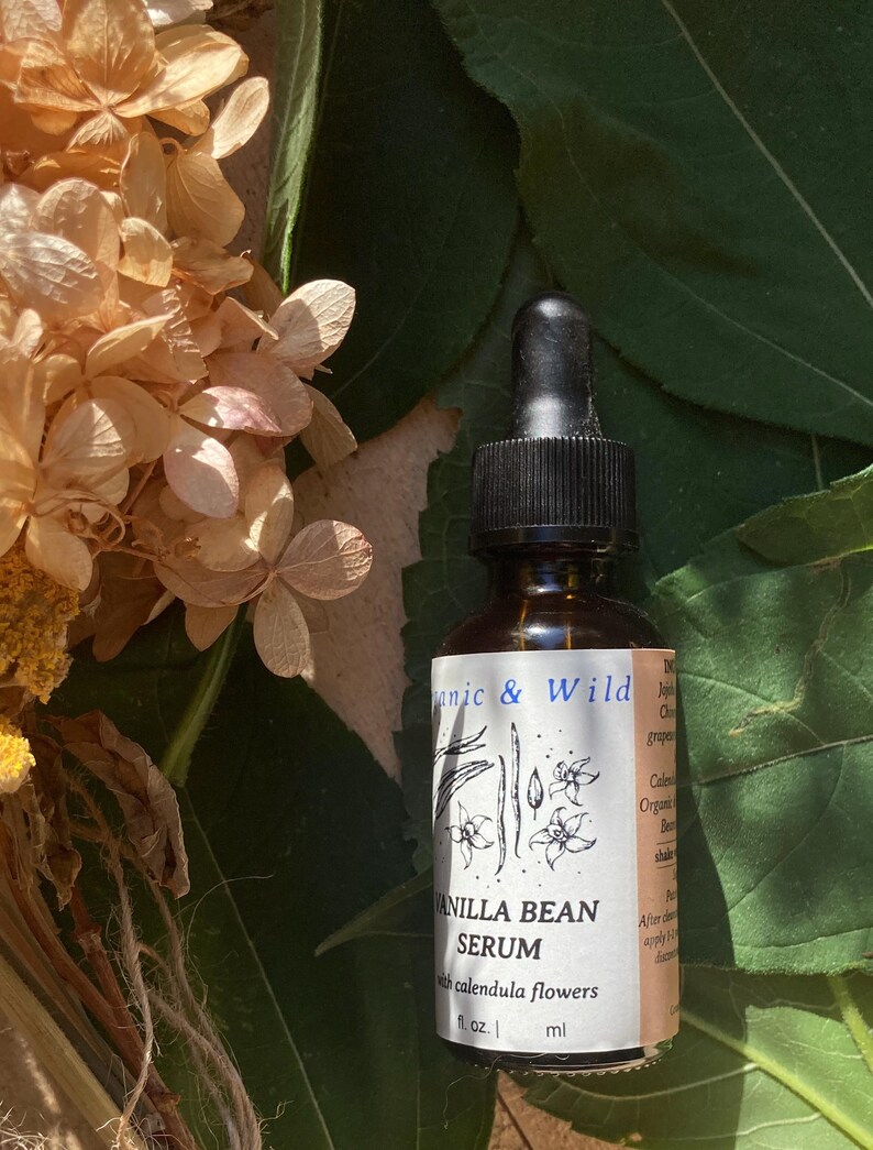 Może przedstawiać: Brązowa szklana butelka serum waniliowego z czarną pipetą, na tle zielonych liści i suszonych kwiat&oacute;w. Etykieta głosi "Organic & Wild" i "Vanilla Bean Serum with calendula flowers."