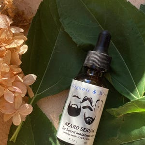 Może przedstawiać: Brązowa szklana butelka serum do brody z czarną pipetą, z napisem "Organic & Wild". Etykieta zawiera ilustracje brod i wąs&oacute;w. Butelka leży na podłożu z zielonych liści i suszonych kwiat&oacute;w.