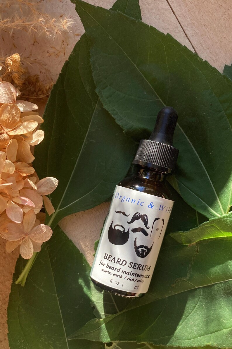 Może przedstawiać: Brązowa szklana butelka serum do brody z czarną pipetą, z napisem "Organic & Wild". Etykieta zawiera ilustracje brod i wąs&oacute;w. Butelka leży na podłożu z zielonych liści i suszonych kwiat&oacute;w.