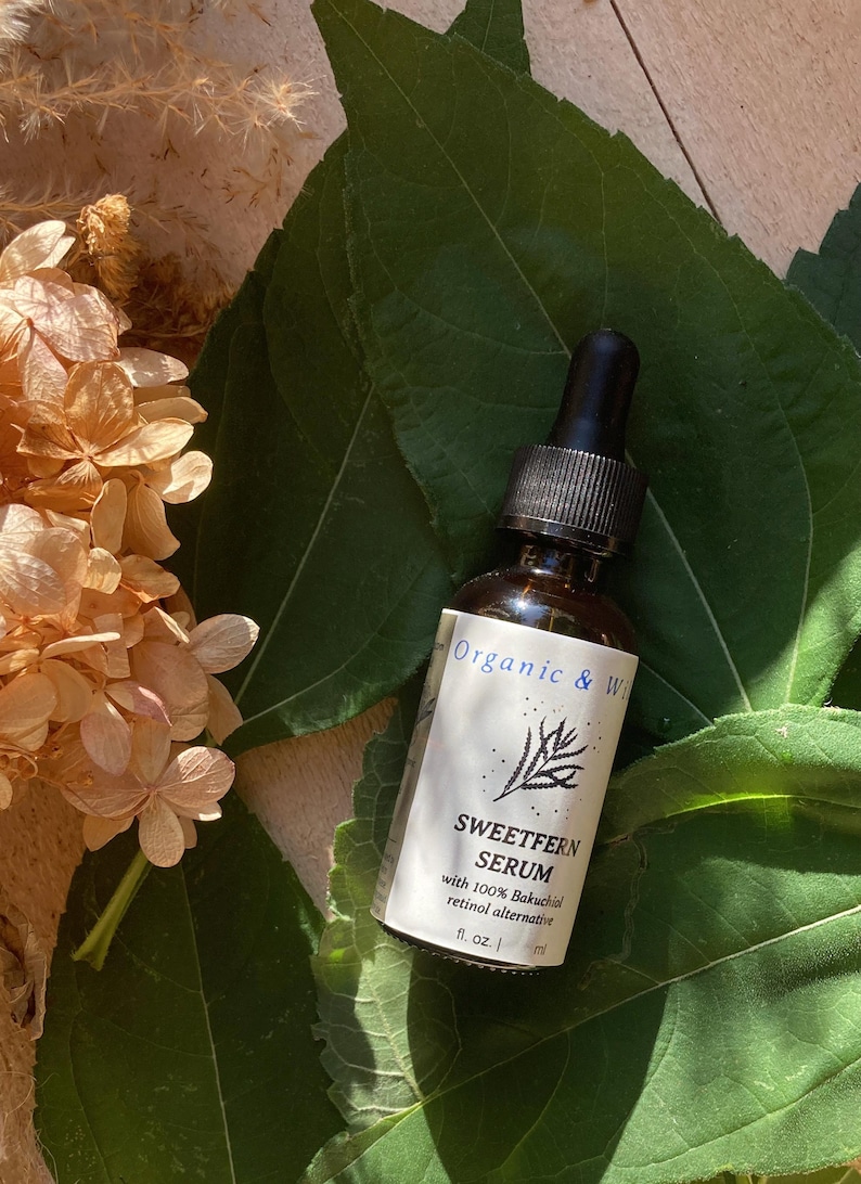 Może przedstawiać: Brązowa szklana butelka Sweetfern Serum z czarną pipetą, na tle zielonych liści i suszonych kwiat&oacute;w. Etykieta głosi "Organic & Wild" i "Sweetfern Serum with 100% Bakuchiol retinol alternative".