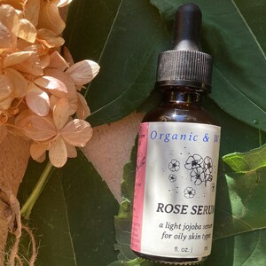 Może przedstawiać: Brązowa szklana butelka z czarnym zakraplaczem, oznaczona jako "ROSE SERUM", lekkie serum jojoba do cery tłustej. Butelka stoi na zielonych liściach, z suszonymi beżowymi kwiatami w tle.