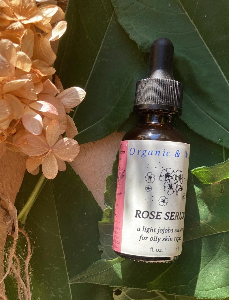 Może przedstawiać: Brązowa szklana butelka z czarnym zakraplaczem, oznaczona jako "ROSE SERUM", lekkie serum jojoba do cery tłustej. Butelka stoi na zielonych liściach, z suszonymi beżowymi kwiatami w tle.