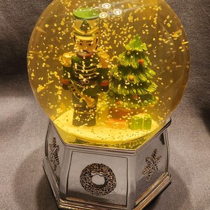May include: A Christmas snow globe featuring a toy soldier and a Christmas tree inside a clear globe filled with yellow liquid and glitter. The base is silver with snowflake and wreath designs.