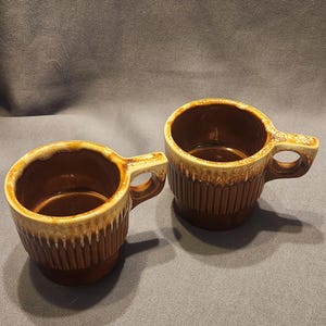 May include: Two brown ceramic mugs with a textured, ribbed design. The rims and handles have a lighter, speckled glaze. The mugs are designed with a handle and are suitable for coffee or tea.